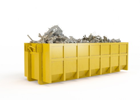 A large, rectangular skip with a bright yellow exterior, constructed from metal panels with visible seams and reinforced edges, filled with a jumble of construction debris including broken concrete blocks, bricks, and rubble. The debris appears rough in texture, with some pieces showing jagged edges and dusty surfaces. The skip is positioned on a clean, white surface with a plain white background, emphasizing the industrial nature of the waste collection. The yellow skip is situated in a way that suggests it is awaiting transport or has recently been filled, relevant to commercial waste removal services in the Pinner area. The overall scene depicts a typical waste management scenario, supporting the services offered by Commercial Waste Removal Pinner for business collections of construction and general waste materials.