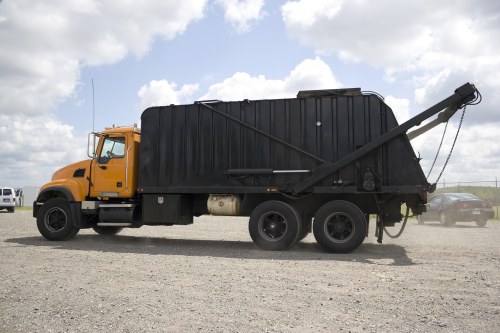 A large commercial garbage collection truck parked on a gravel surface under a partly cloudy sky, with a black waste container mounted on the back, featuring a hydraulic lifting mechanism extended towards the top at an angle. The truck has a yellow cab with a black chassis and six wheels visible, indicating its capacity for industrial rubbish collection. Surrounding the truck, in the background, are several smaller vehicles and open countryside typical of the area, which could be near Pinner in North London. The scene reflects a professional waste management operation, aligned with services offered by Commercial Waste Removal Pinner, focused on business waste collections and rubbish disposal. The vehicle’s textured metal surface, the sturdy rubber tires, and the hydraulic components are clearly visible, supporting the context of an efficient, commercial rubbish removal process, suitable for local business premises or municipal waste services in the area.