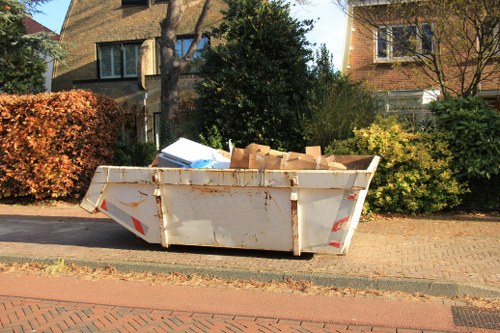 A large, rectangular metal skip with a weathered, off-white finish and visible rust stains is positioned on a paved driveway beside a residential street. The skip is filled with an assortment of brown cardboard boxes, some torn and crumpled, with their flaps open and slightly leaning. Surrounding the skip are green bushes and small trees, with a background of brick and stone houses featuring pitched roofs, large windows, and chimney stacks. The street is lined with a low brick wall and a narrow pavement, typical of suburban areas within the vicinity of Pinner. The scene is illuminated by natural daylight, creating soft shadows and highlighting the textures of the materials. This image is associated with rubbish removal services, emphasizing the collection and disposal of bulky waste, and visualizes an environment where Commercial Waste Removal Pinner operates to support local businesses and residents with waste clearance.